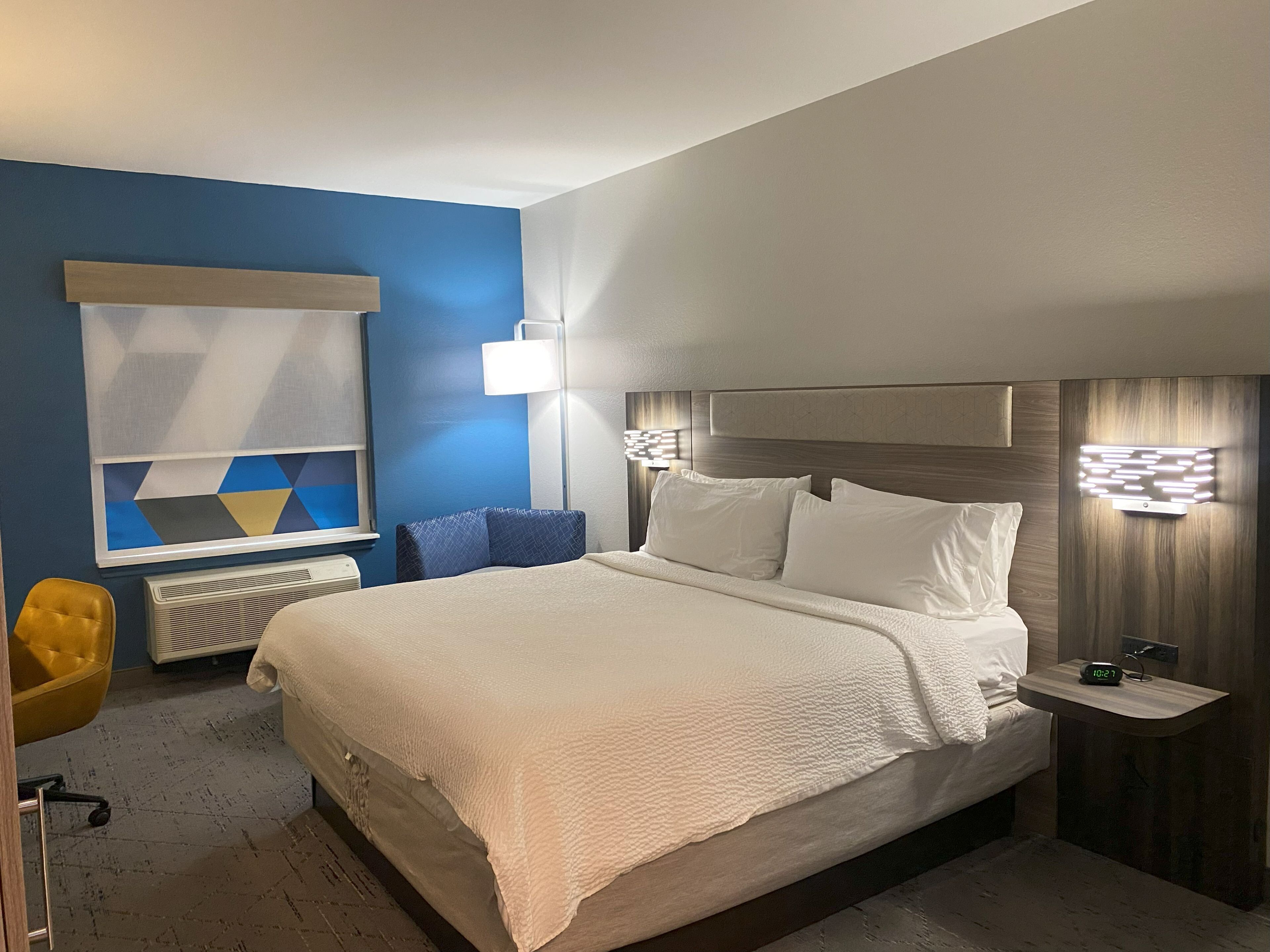 A hotel room with a large bed, a desk, a chair, a radiator, and a wall-mounted lamp.
