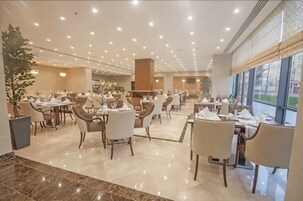 Un comedor de hotel espacioso con varias mesas y sillas, una planta grande en maceta y vistas a través de amplios ventanales.