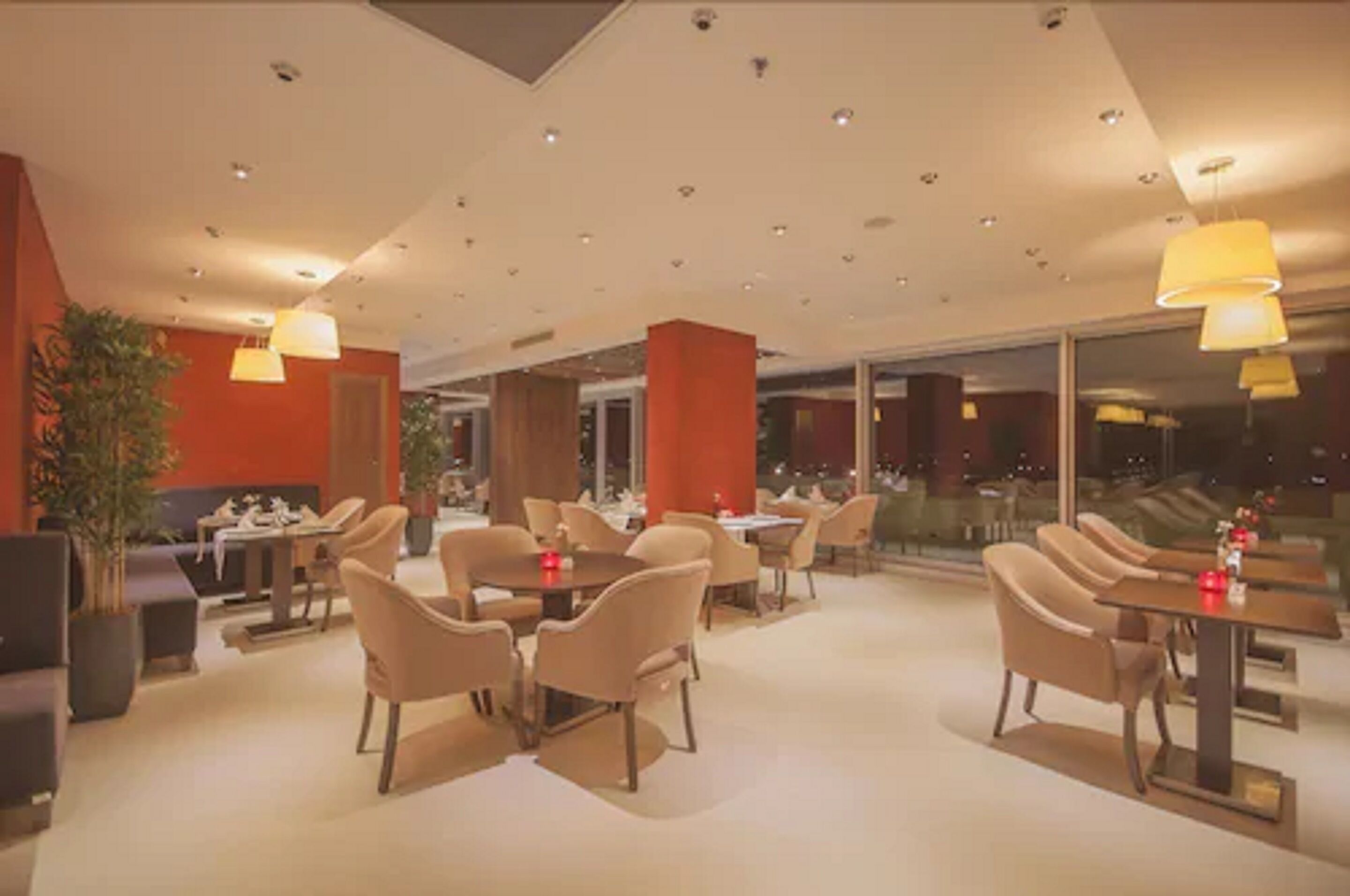 Un restaurante moderno con mesas redondas y sillas color beige, que presenta una paleta de colores cálidos y ventanas amplias.