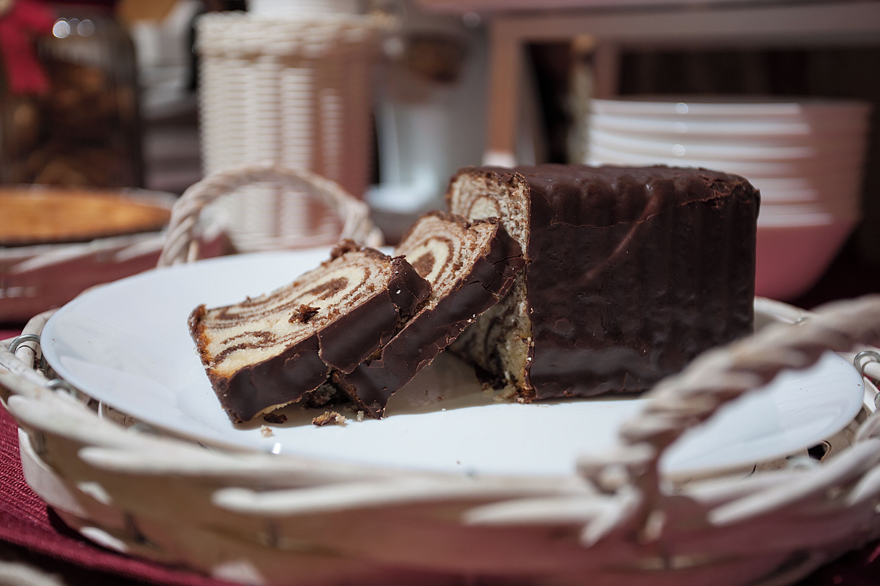 A slice of chocolate-covered cake on a white plate with a wicker basket in the background.