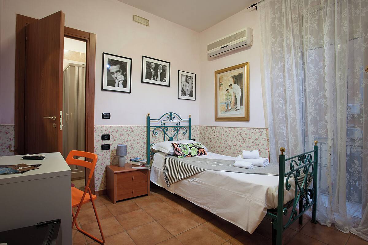 A bedroom with a bed, a desk, a chair, a nightstand, and a wall with framed pictures.