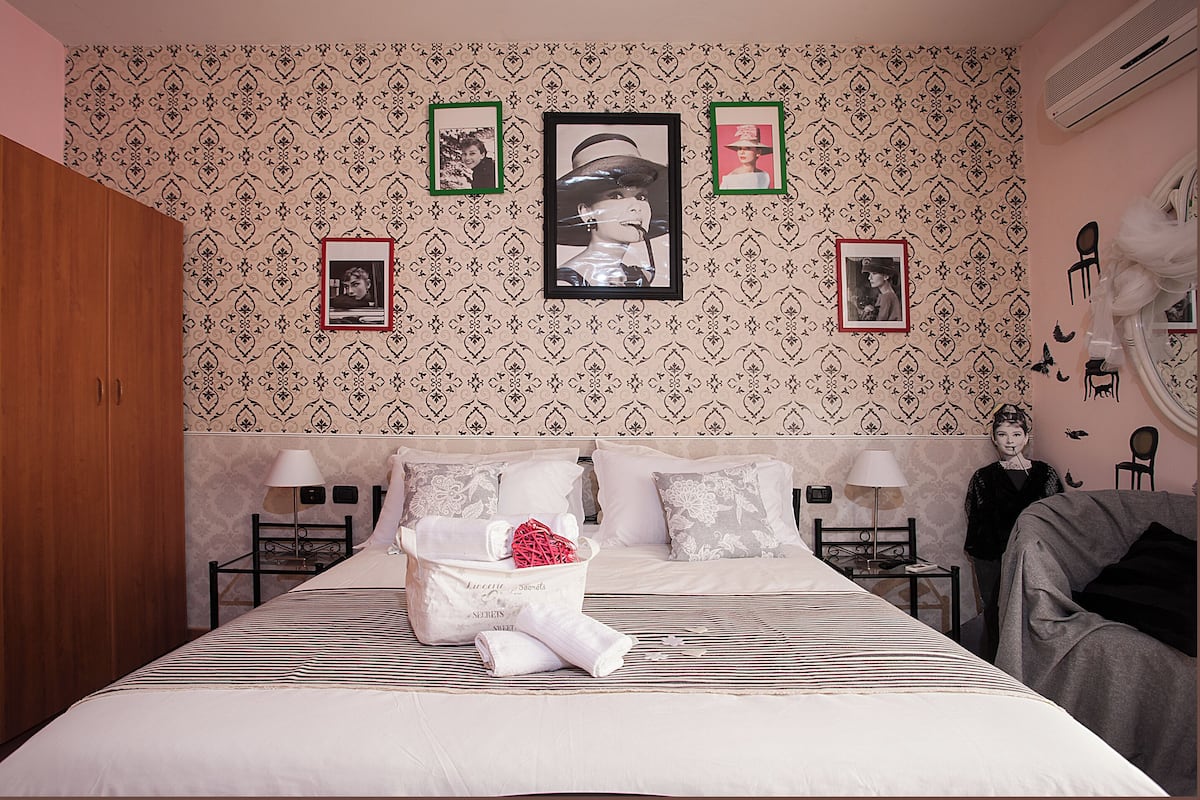 A hotel room with a bed, a nightstand, a chair, and a wall with framed pictures.
