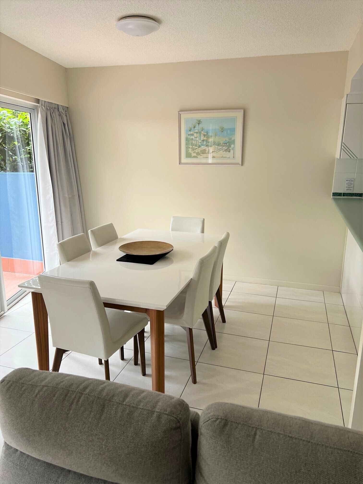 A dining area with a white table and chairs, a framed picture on the wall, and a view of greenery outside.