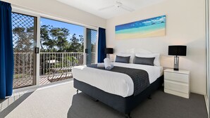 A bedroom with a bed, a nightstand, a ceiling fan, and a painting of a beach. - White Crest Apartments (Torquay)