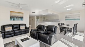 A modern living room with a black leather sofa, a white coffee table, and a dining area with a glass table and black chairs. - White Crest Apartments (Torquay)