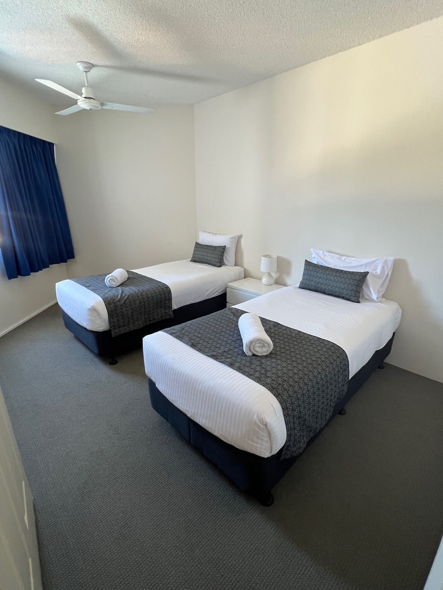 A hotel room with two single beds, a ceiling fan, a nightstand, and a window with curtains.