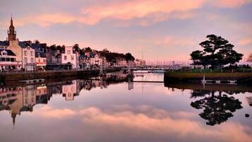 A waterfront town with buildings reflecting in calm water, a prominent church tower, and a large tree.