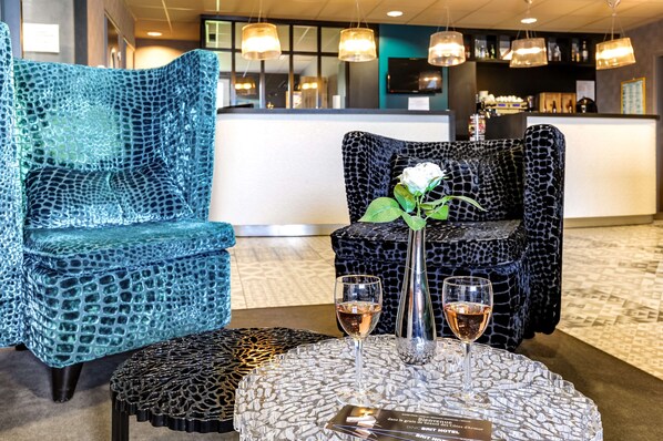 Un área de recepción con dos sillones estampados, una mesita redonda con dos copas de vino y un jarrón con una flor blanca.