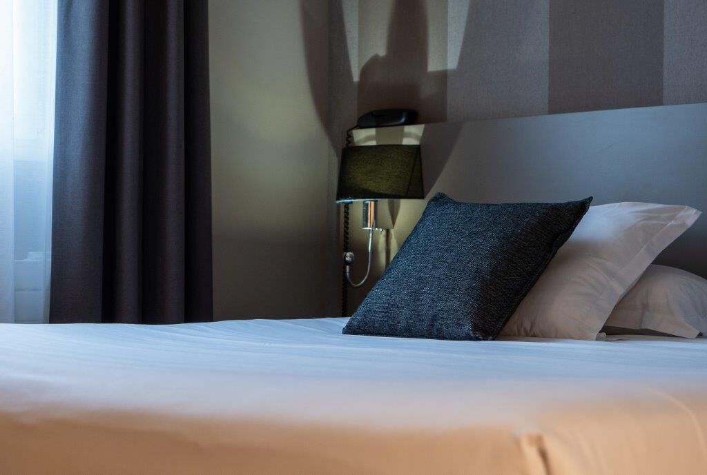 A neatly made bed with white linens, a dark blue pillow, and a bedside lamp with a metallic finish.