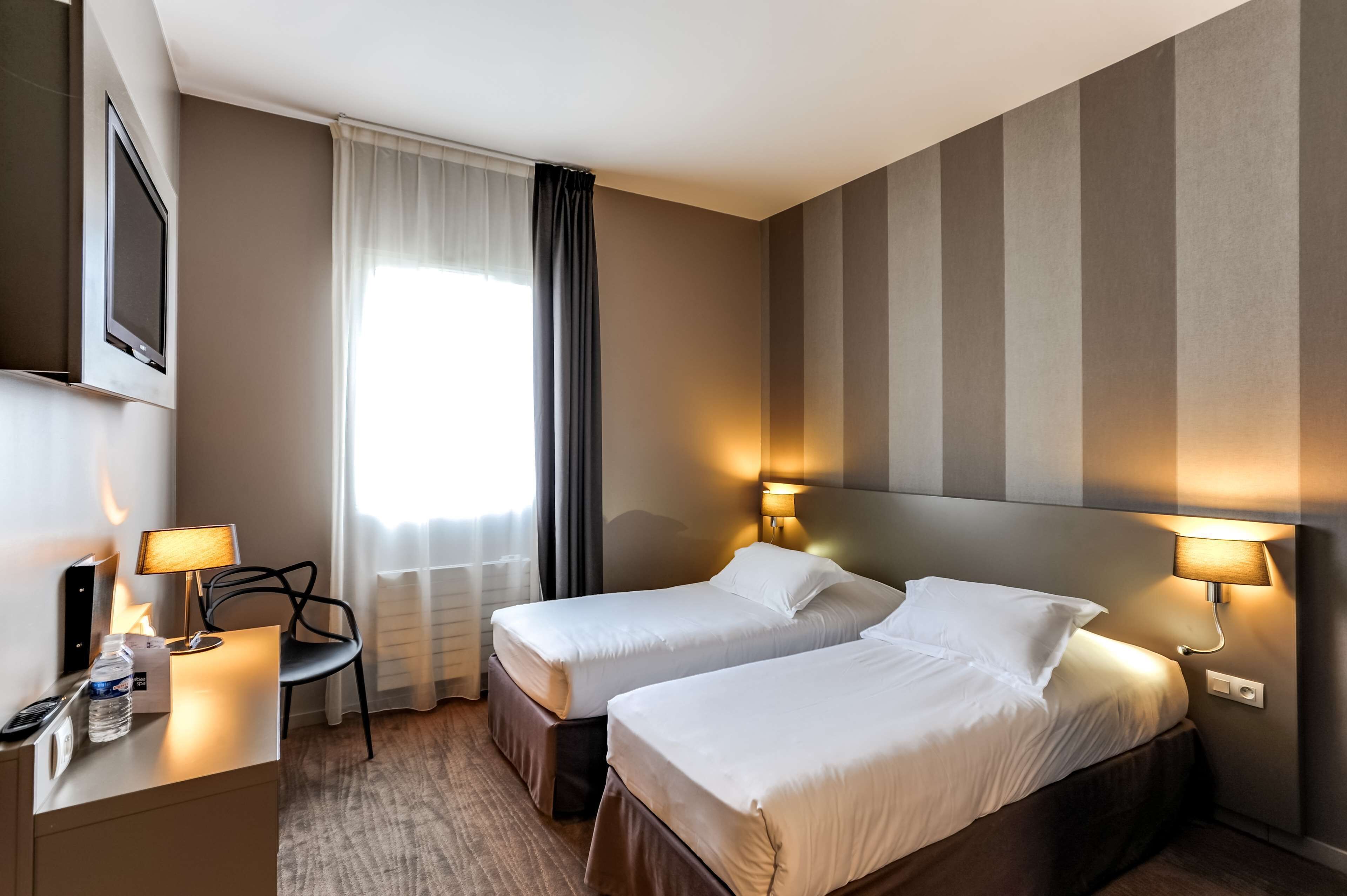 A hotel room with two beds, a desk with a lamp, a chair, and a TV on the wall.