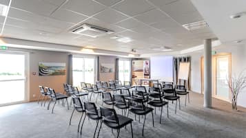 Un salón de conferencias con filas de sillas, ventanas amplias y una pantalla de proyector.