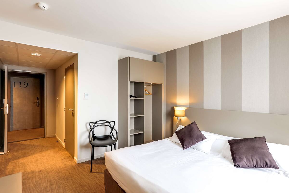 Une chambre d’hôtel avec un lit, une chaise, une armoire et une porte portant le numéro « 119 ».