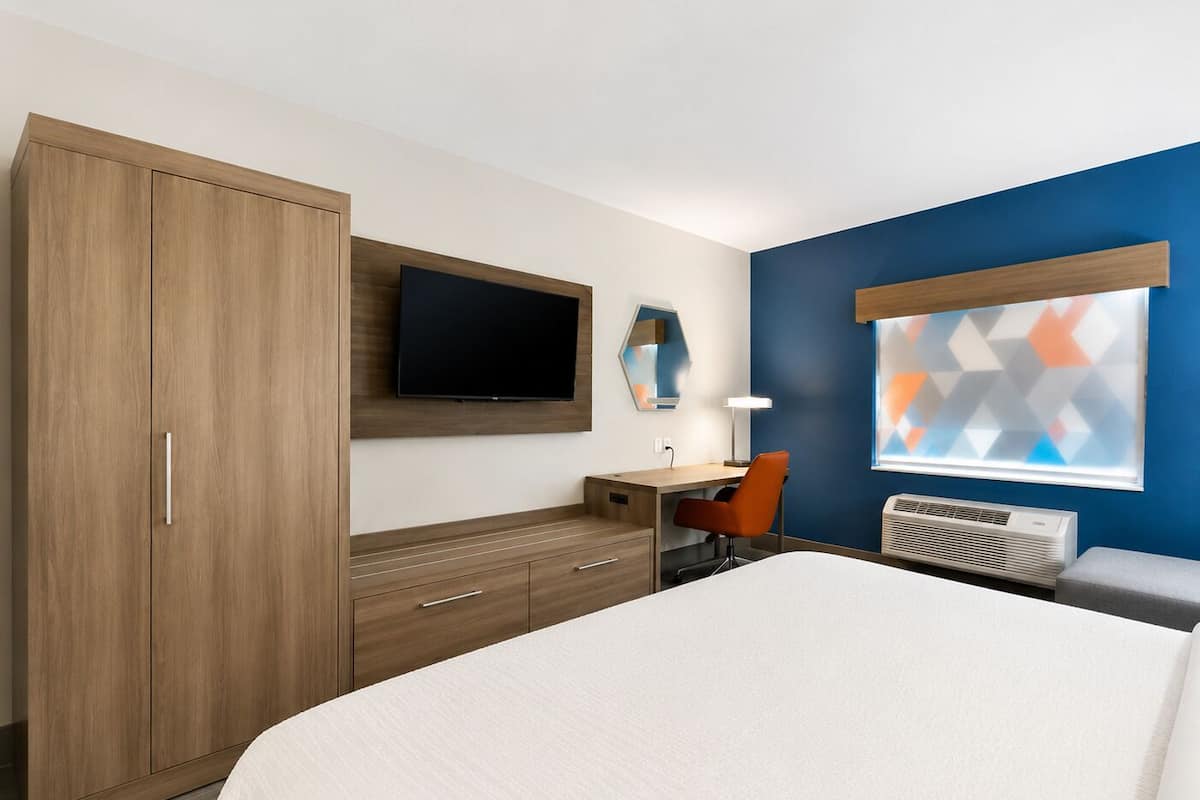 A hotel room with a bed, a desk with a chair, a TV, a heater, and a wardrobe.