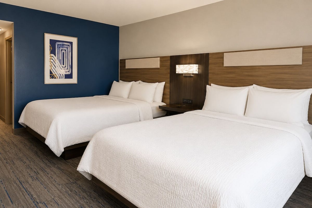 A hotel room with two beds, a blue accent wall, a framed artwork, and a nightstand with a lamp.