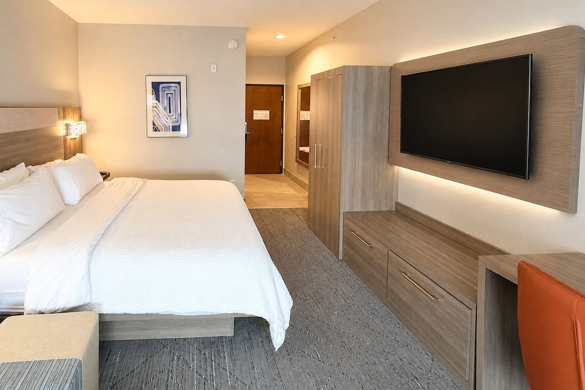 A hotel room with a bed, a flat-screen TV mounted on the wall, a wardrobe, and a desk with a chair.