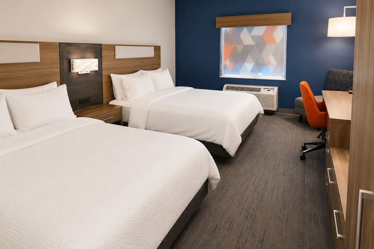 A hotel room with two beds, a desk, a chair, a window with a geometric view, and a wall-mounted air conditioner.