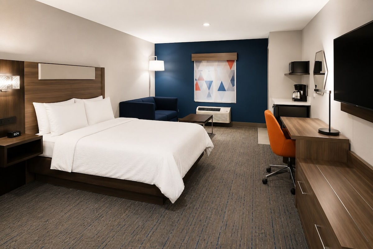 A hotel room with a bed, desk, chair, TV, and artwork on the wall.