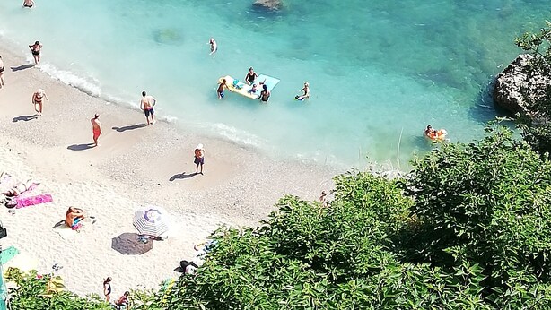 Een strand met helder turquoise water, rotsformaties en mensen die zwemmen en zonnebaden.