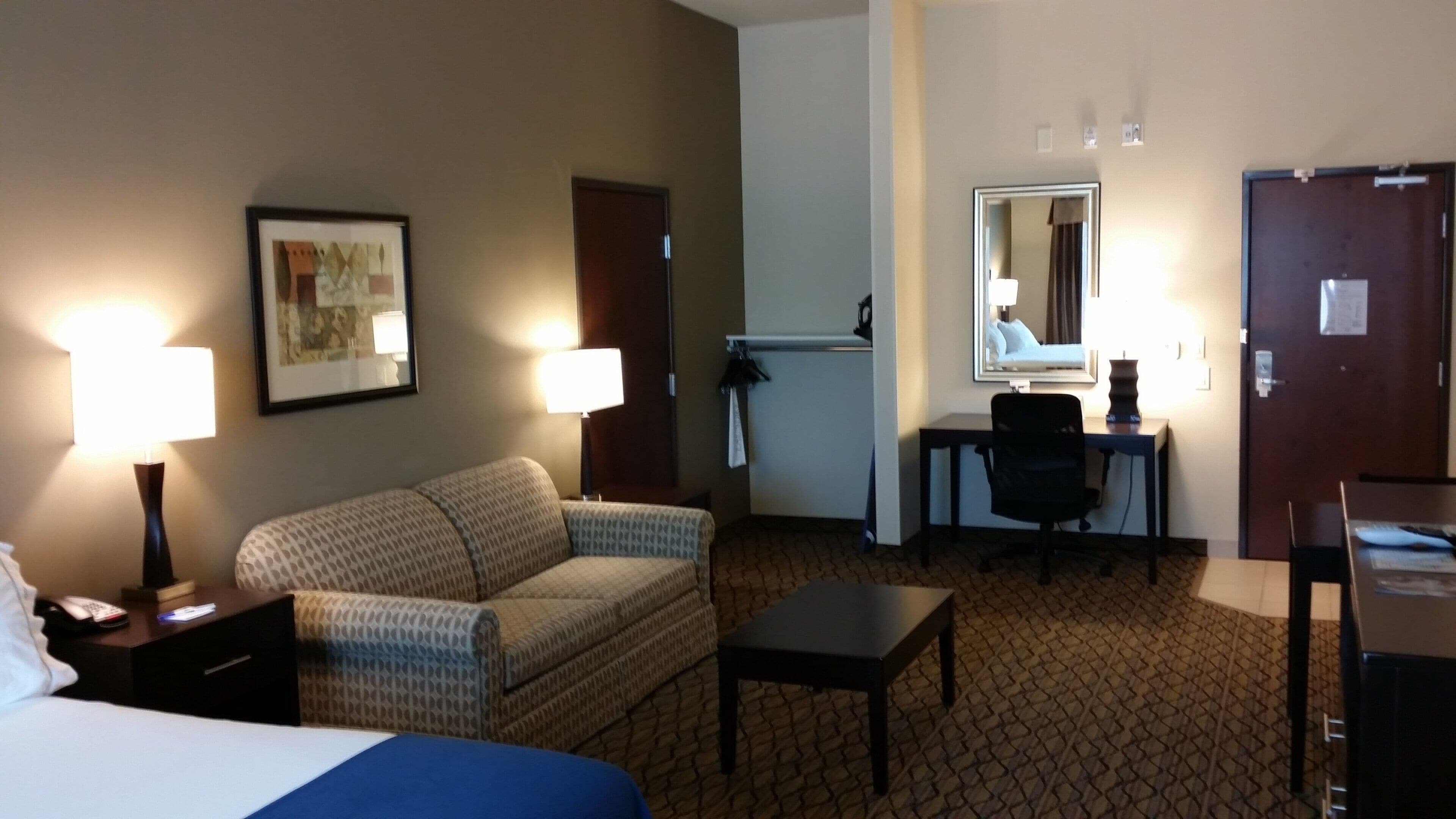 A hotel room with a bed, a sofa, a desk, and a chair. There are lamps on the desk and a framed picture on the wall.
