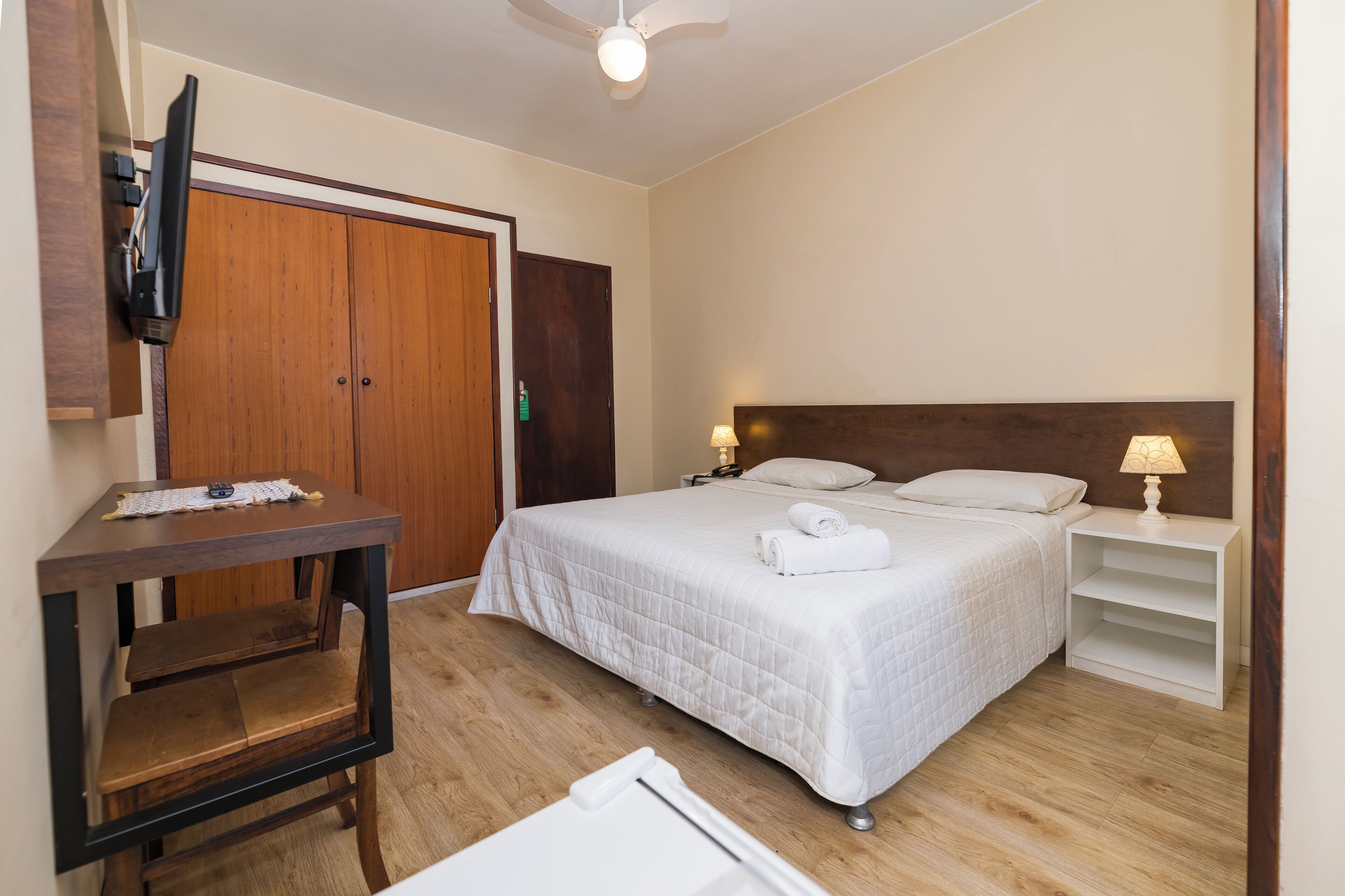 A hotel room with a bed, bedside tables, a TV mounted on the wall, a wooden wardrobe, and a ceiling fan.