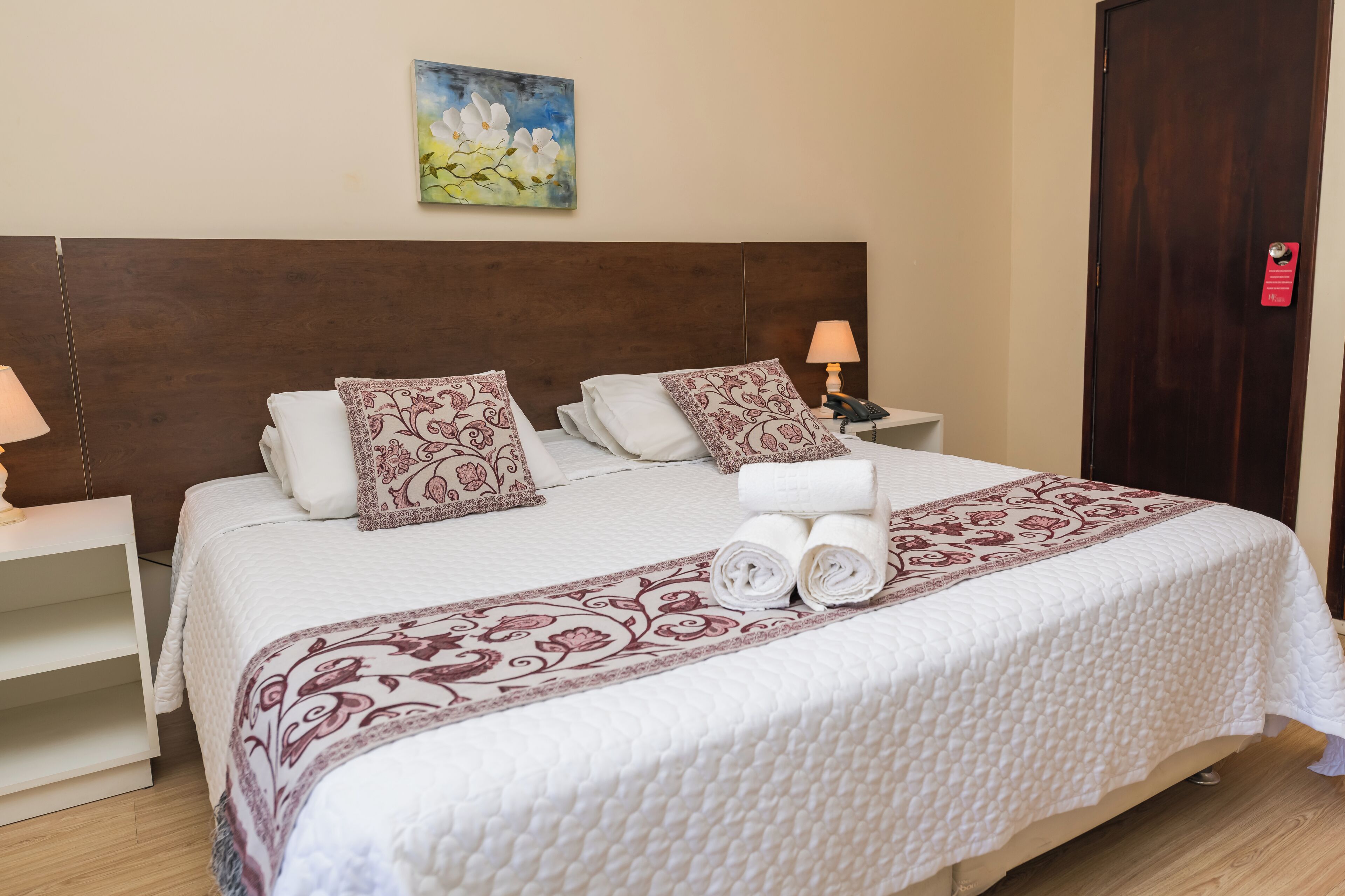 A neatly made bed with a floral-patterned bedspread and pillows, a bedside lamp, and a painting on the wall.