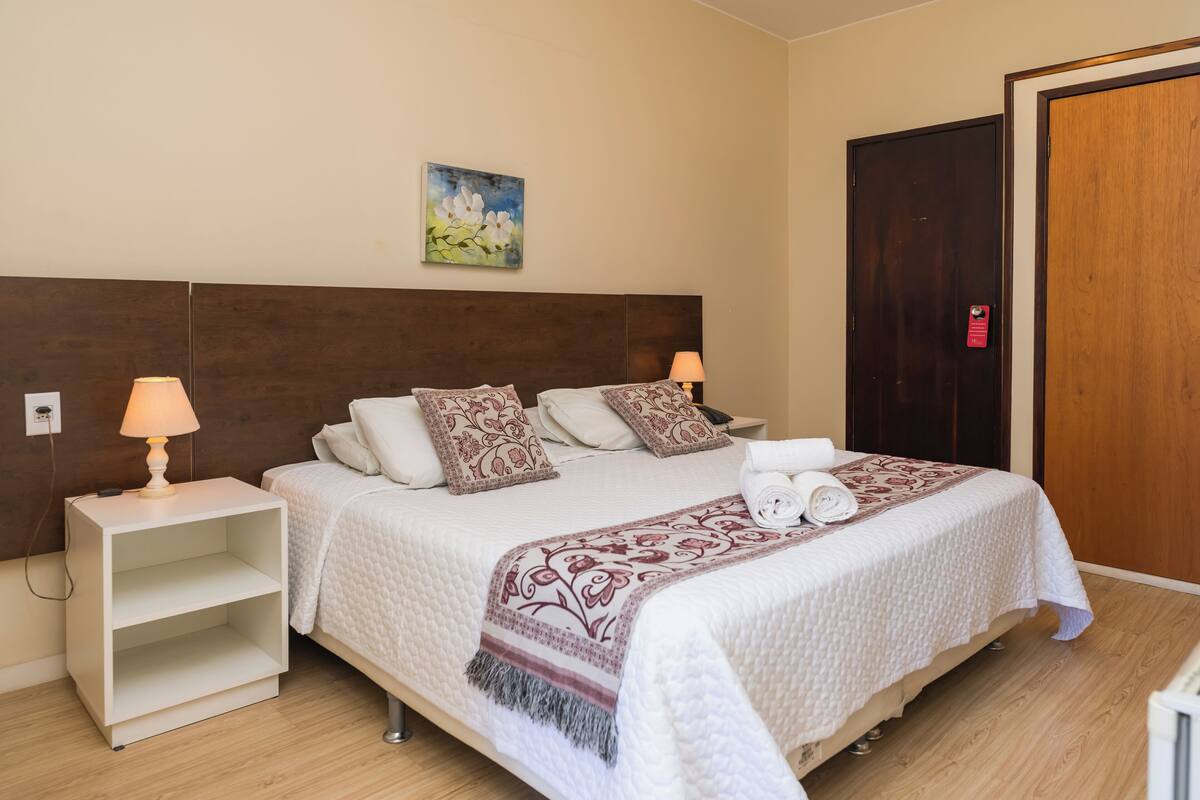 A neatly arranged bedroom with a large bed, two bedside tables, a nightstand, a wooden door, and a wall-mounted painting.