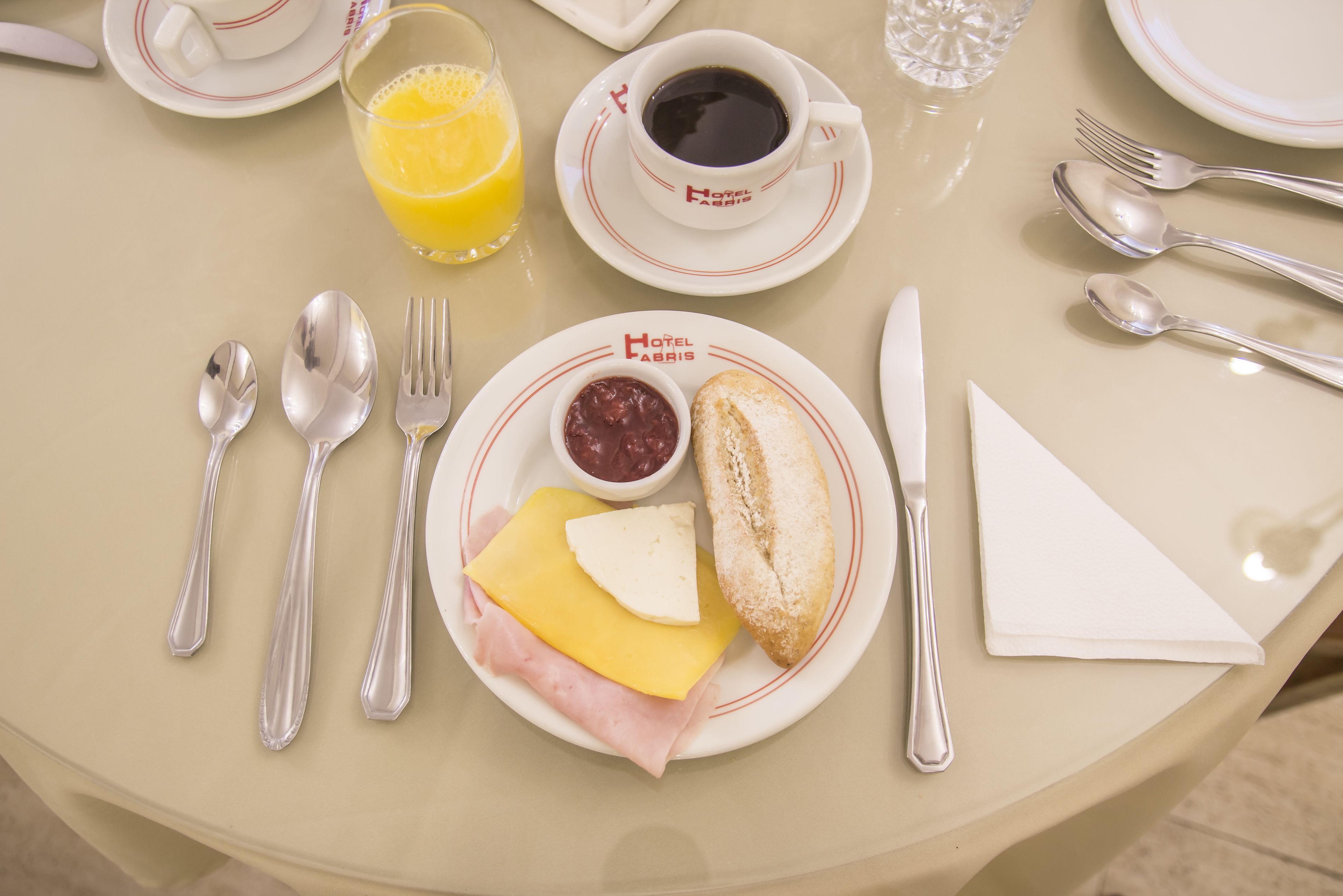 Una mesa color beige con un plato de jamón, queso y pan, un vaso de jugo de naranja y una taza de café.