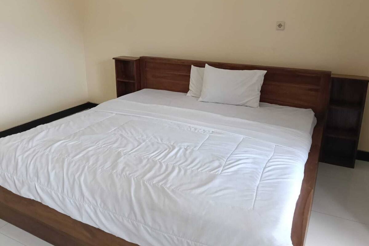 Ein ordentlich bezogenes Bett mit Holz-Kopfteil und weißer Bettwäsche in einem schlichten Zimmer mit schlichten Wänden.
