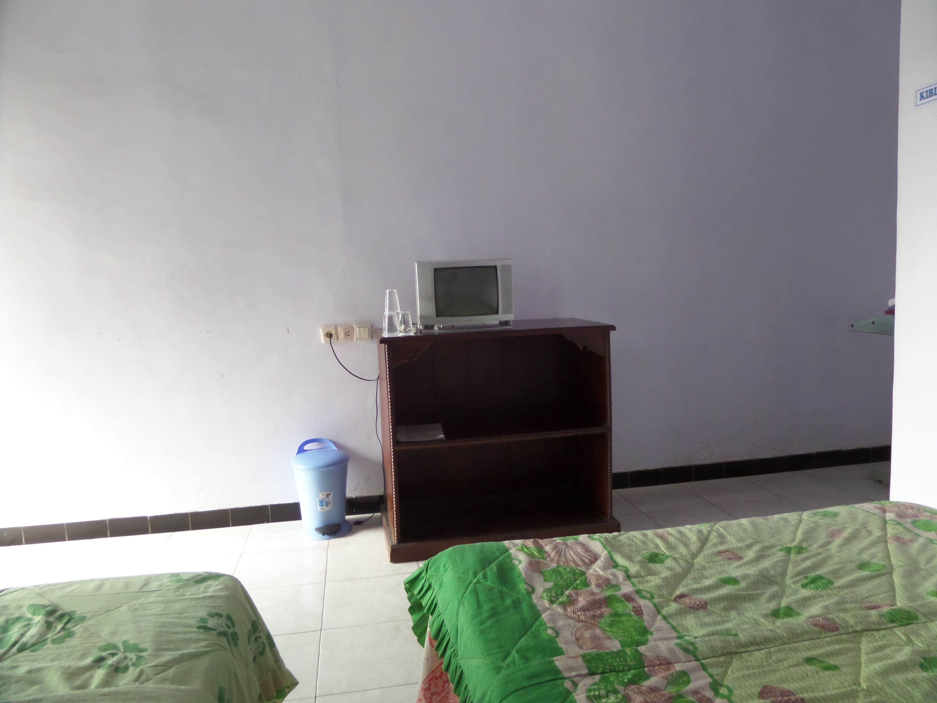 Un dormitorio con una cama, un pequeño estante de madera con un televisor y un cubo de basura azul.