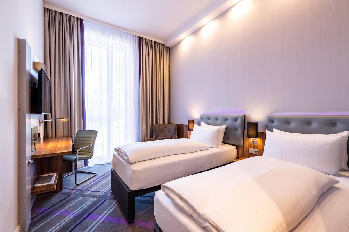 Ein Hotelzimmer mit zwei Betten, einem Schreibtisch, einem Stuhl, einem Fernseher und einem großen Fenster mit Vorhängen.