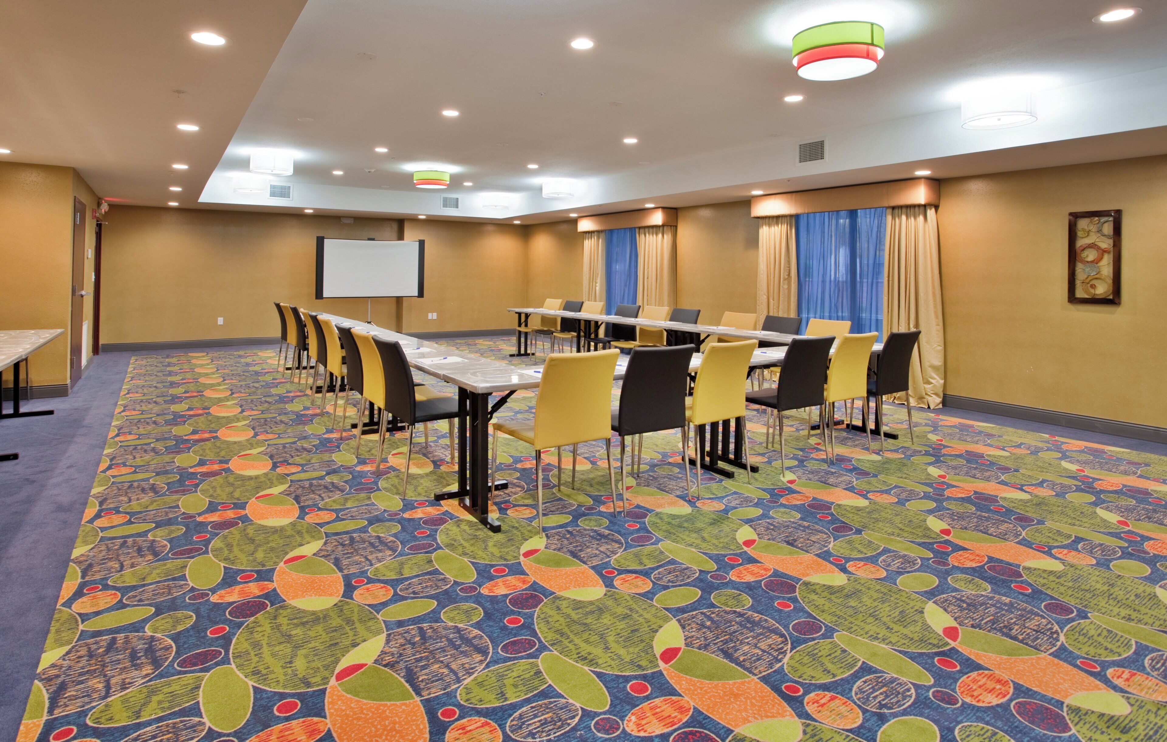 A conference room with a long table, chairs, a projector screen, and a colorful carpet.