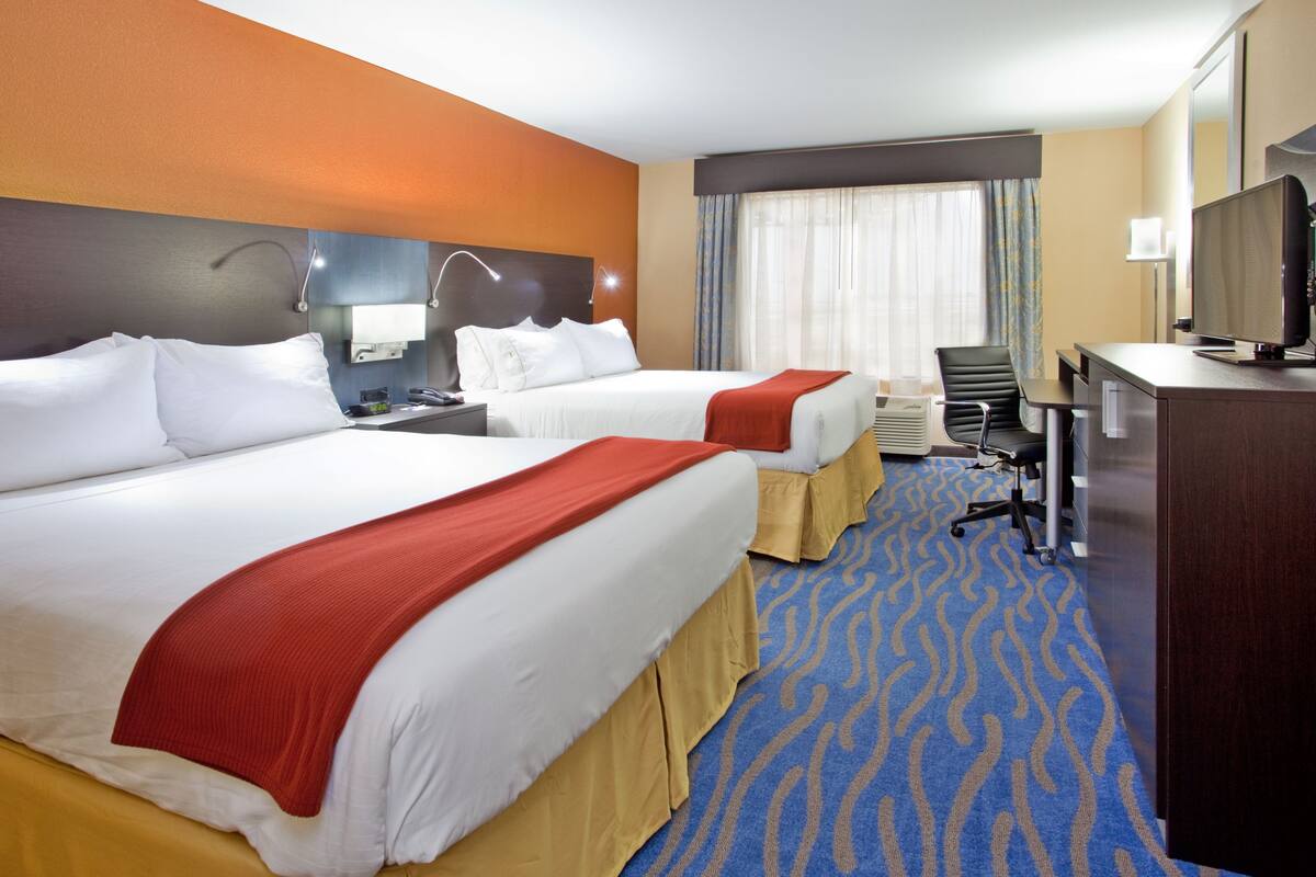 A hotel room with two beds, a desk, a television, and a large window with curtains.