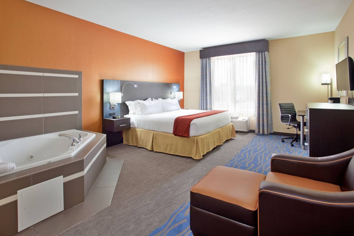 A hotel room with a large bed, a separate bathroom with a bathtub, a desk with a chair, and a large window with curtains.