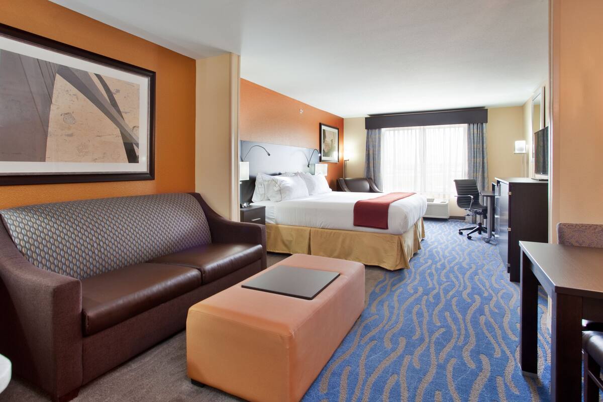 A hotel room with a brown leather sofa, a bed with a red blanket, a desk with a computer, and a large framed picture on the wall.