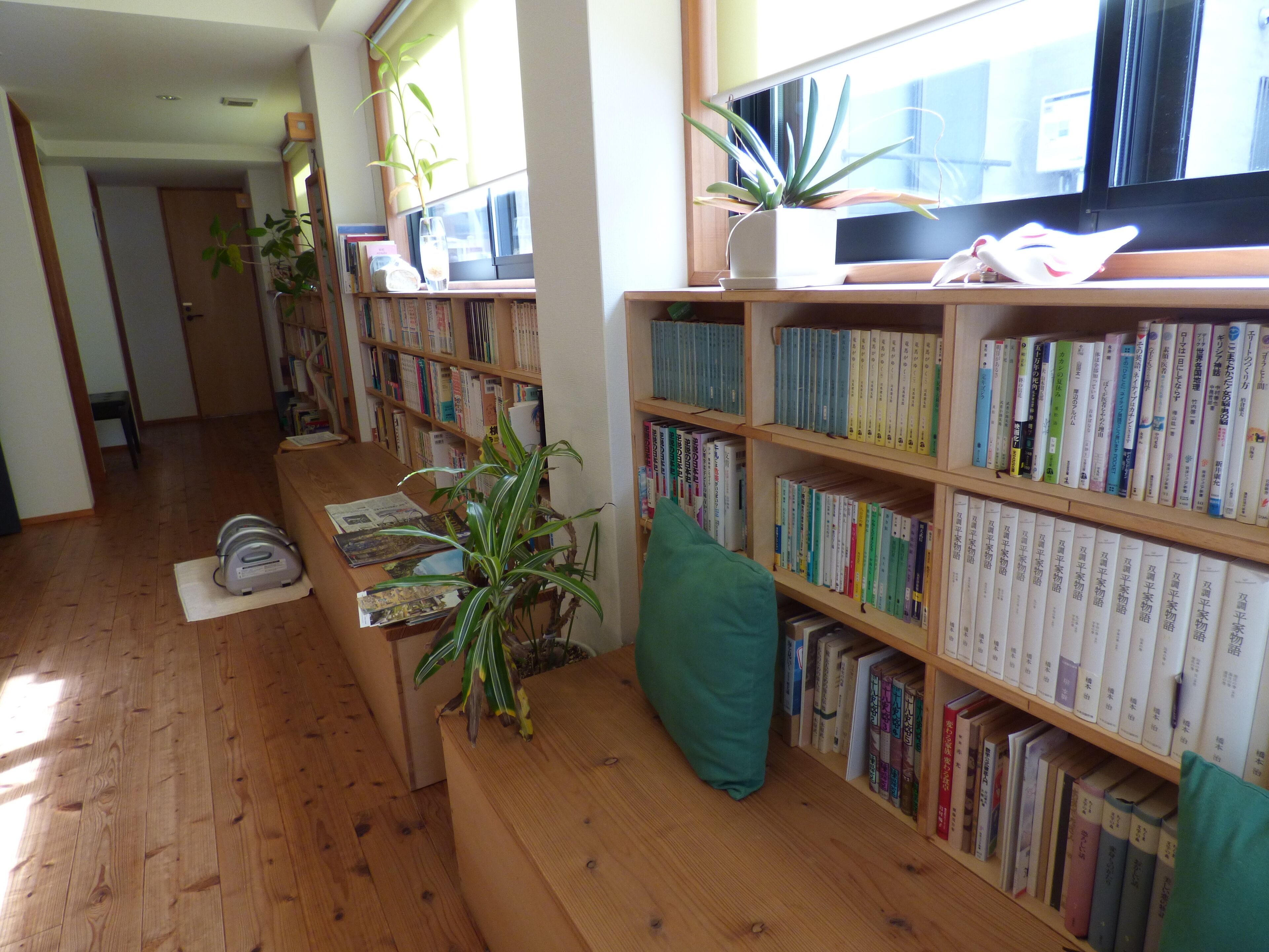 A library with wooden bookshelves filled with books, a green cushioned bench, and potted plants.