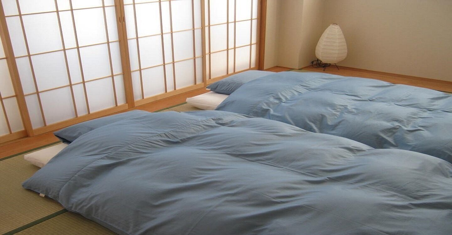 Une pièce avec un sol recouvert de tatamis, un paravent en papier de riz (shoji) et deux lits avec une literie bleue.