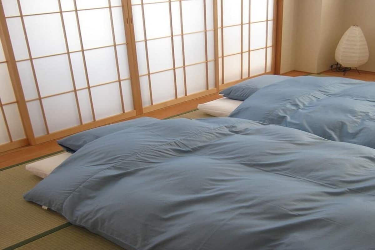 Une pièce avec un sol recouvert de tatamis, un paravent en papier de riz (shoji) et deux lits avec une literie bleue.