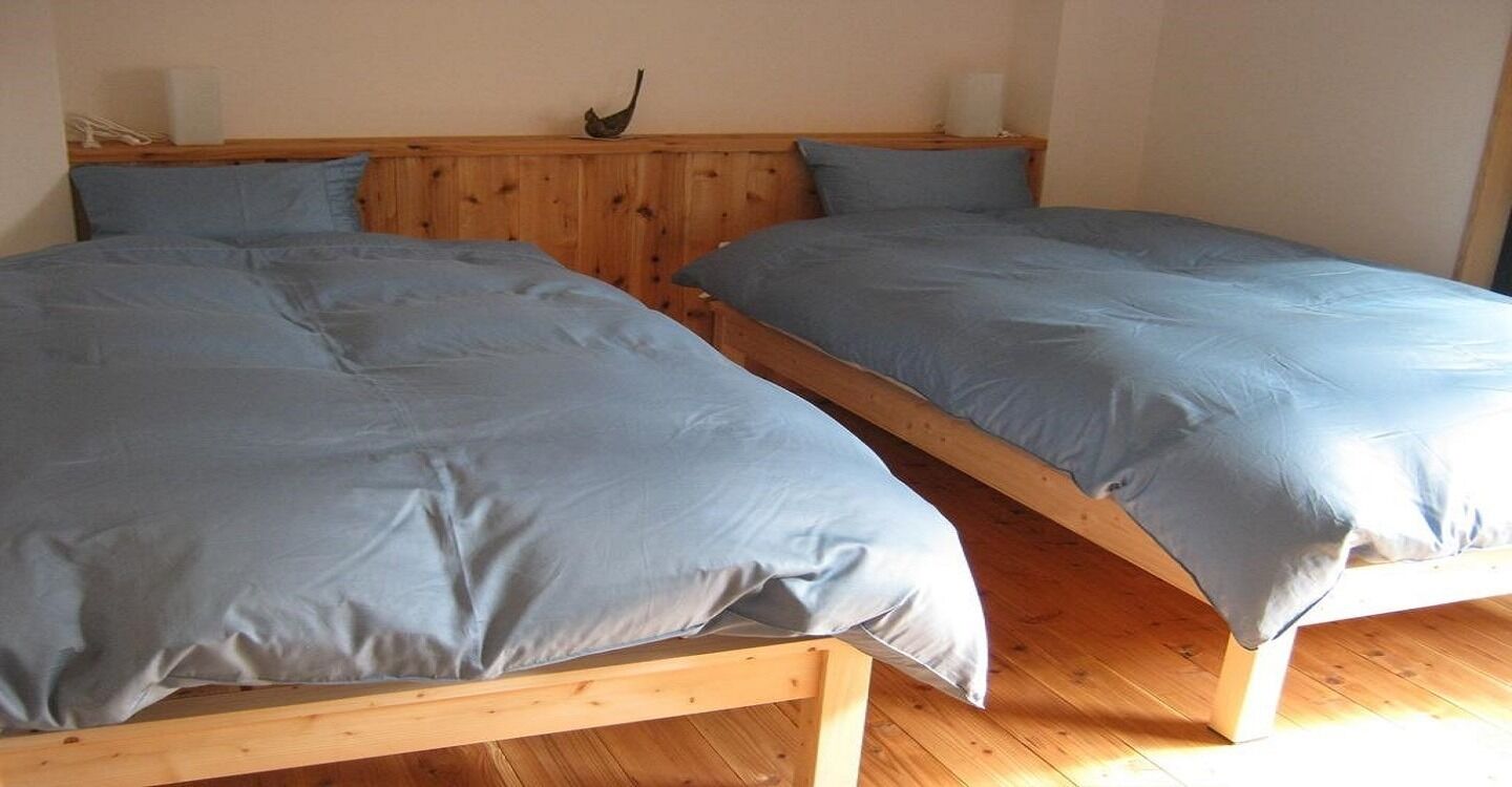 Deux lits en bois, recouverts de draps gris, dans une pièce dont le sol et la tête de lit sont également en bois.
