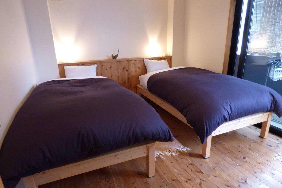 A room with two wooden beds, each with a dark-colored mattress and white pillows. The room has wooden flooring and a large window with blinds.