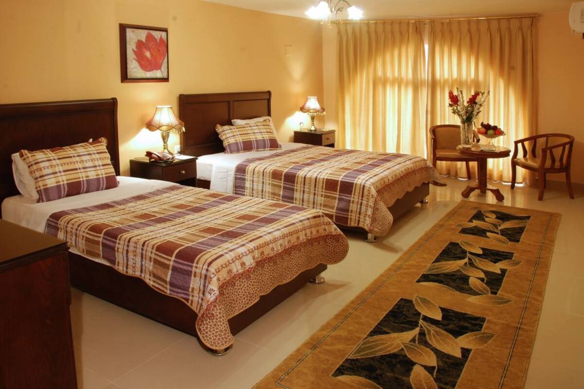 A hotel room with two beds, a wooden headboard, a small table with a lamp, a chair, and a decorative rug.