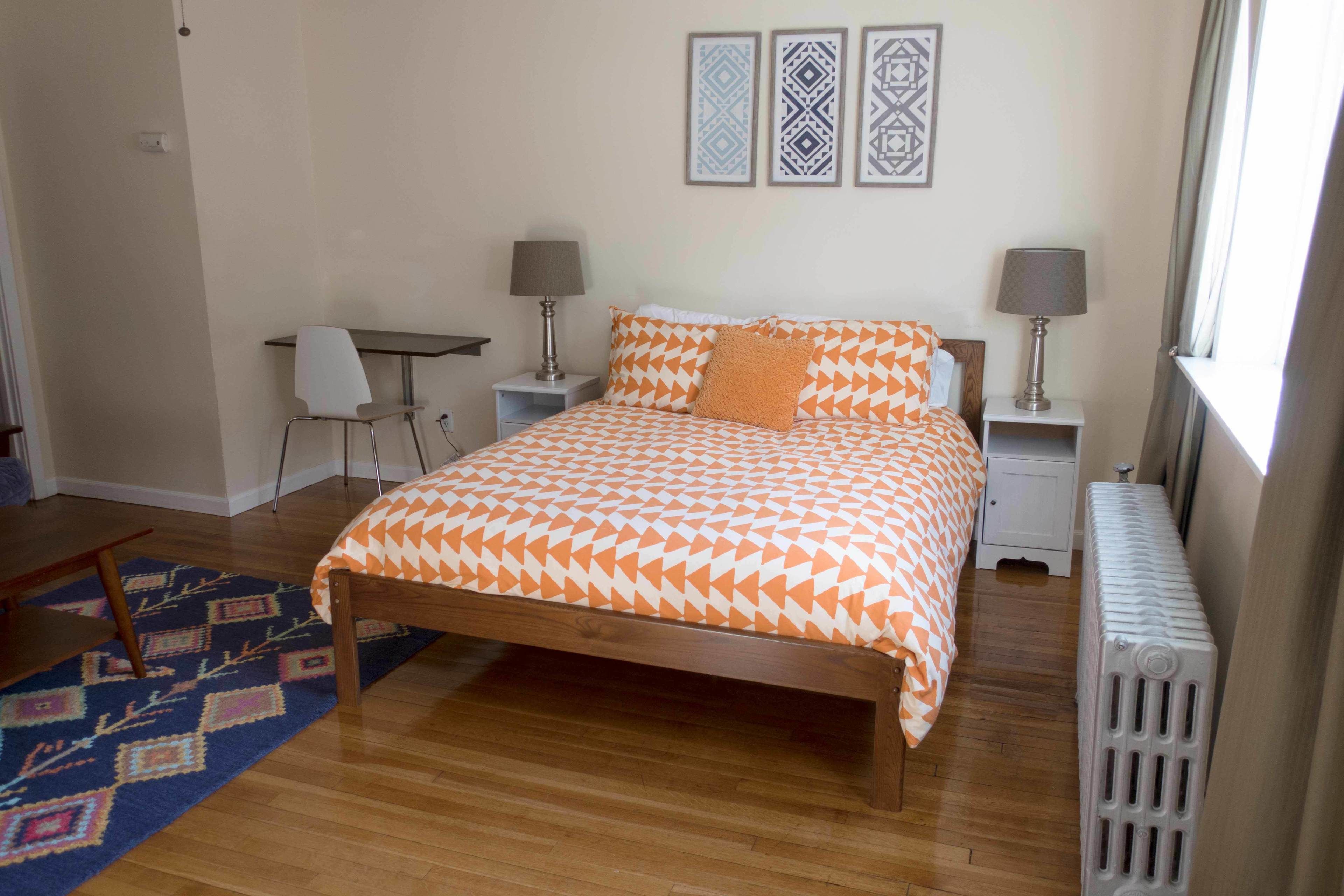Un dormitorio con una cama de madera, dos mesitas de noche con lámparas, un escritorio con una silla, alfombras estampadas y decoración mural.