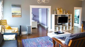 A living room with a sofa, a coffee table, a TV, and a chandelier. There is a radiator and a door leading to another room. - Sauerdough Lodging (Seward)