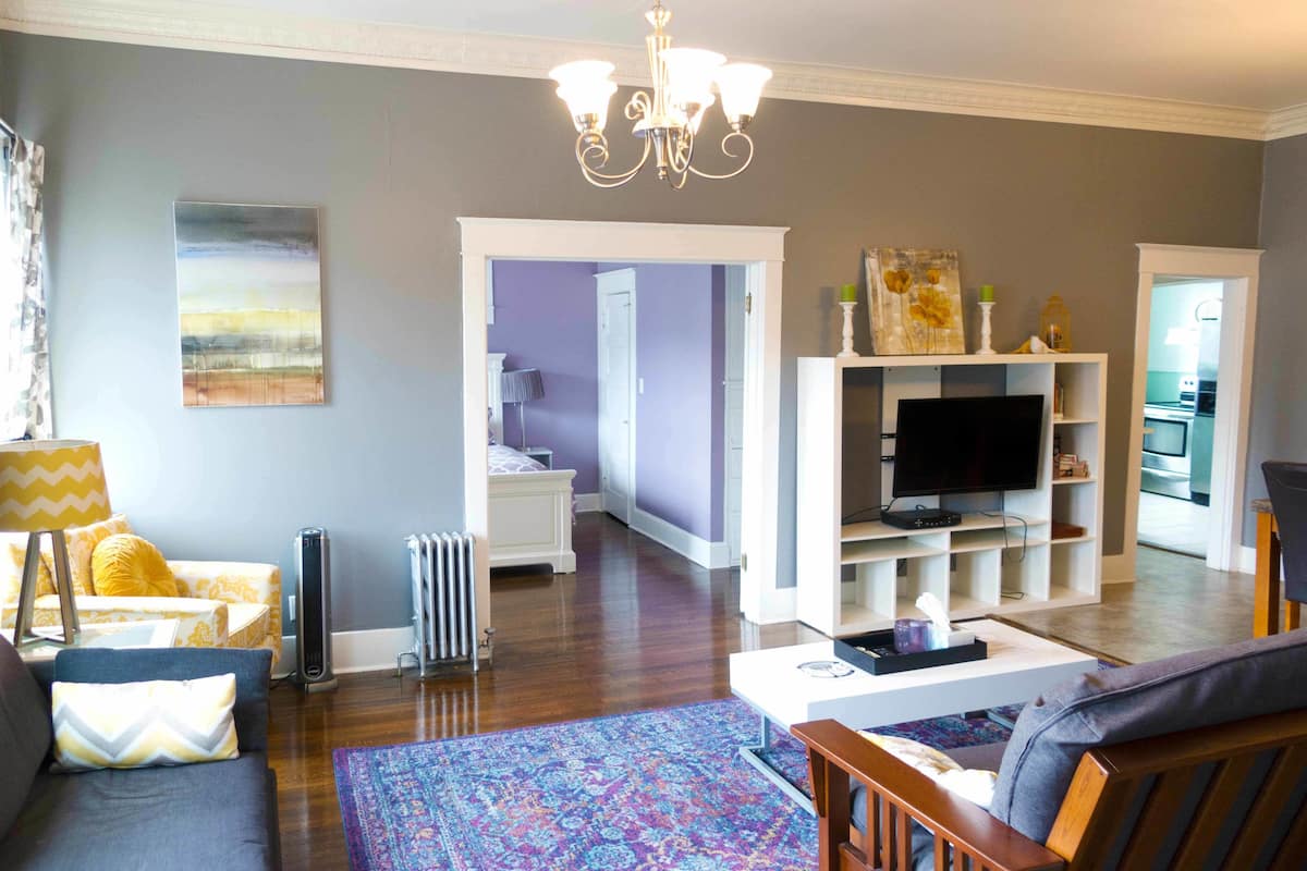 A living room with a sofa, a coffee table, a TV, and a chandelier. There is a radiator and a door leading to another room.