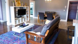A modern living room with a flat-screen TV, a sofa, a dining table with chairs, and a chandelier. - Sauerdough Lodging (Seward)