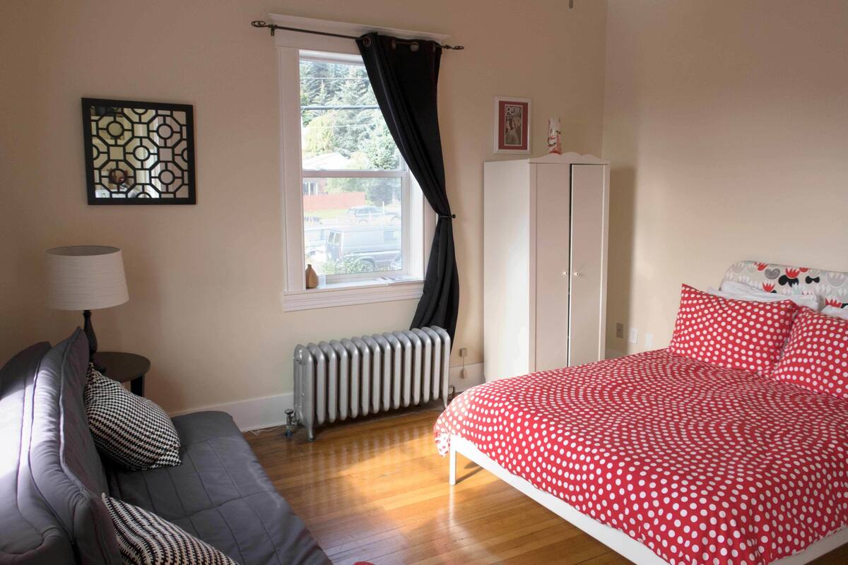 A bedroom with a grey sofa, a red polka dot bedspread, a radiator, and a window with black curtains.