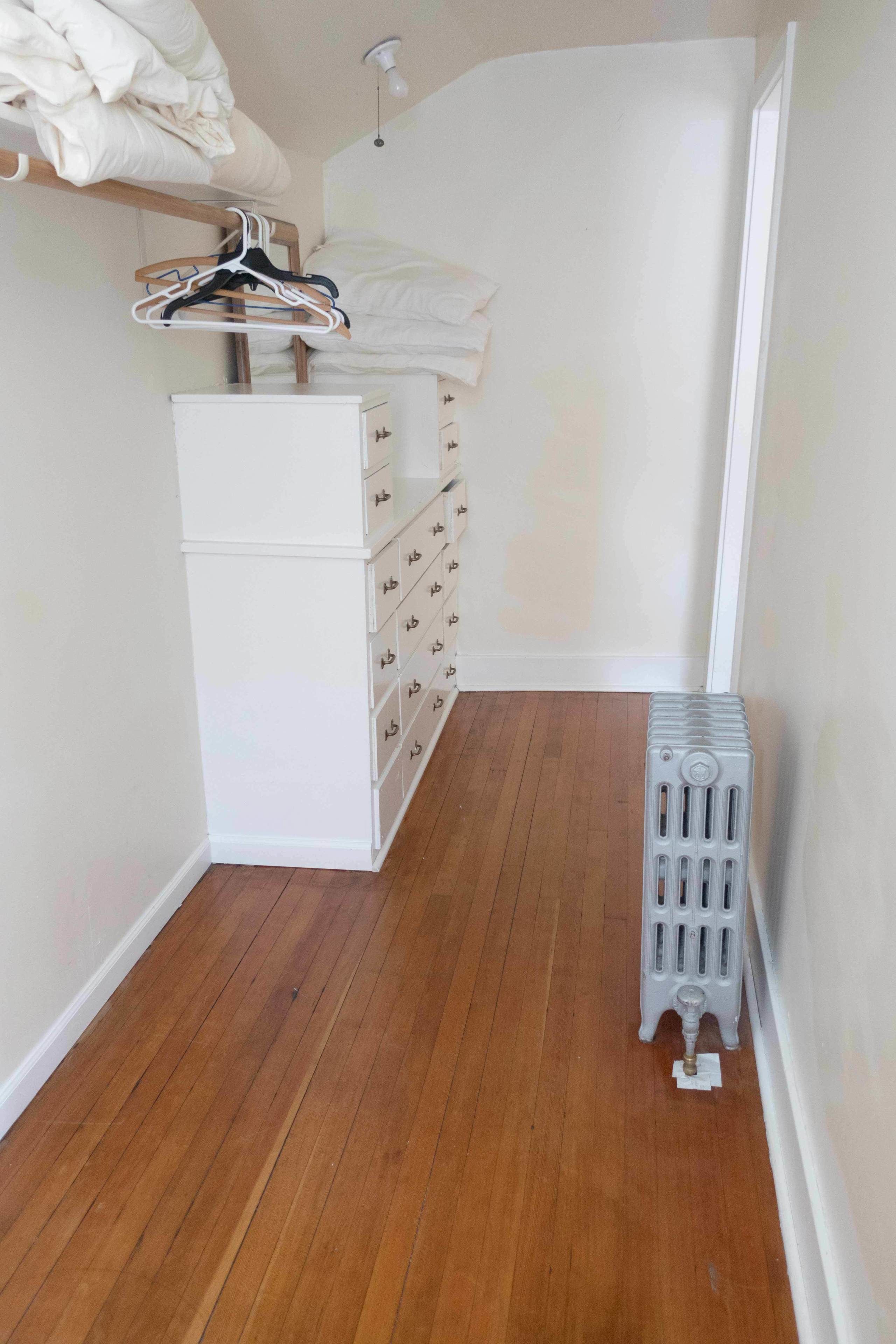Un couloir étroit au plancher en bois, une commode blanche avec de nombreux tiroirs, un radiateur et un portant à vêtements avec des cintres.