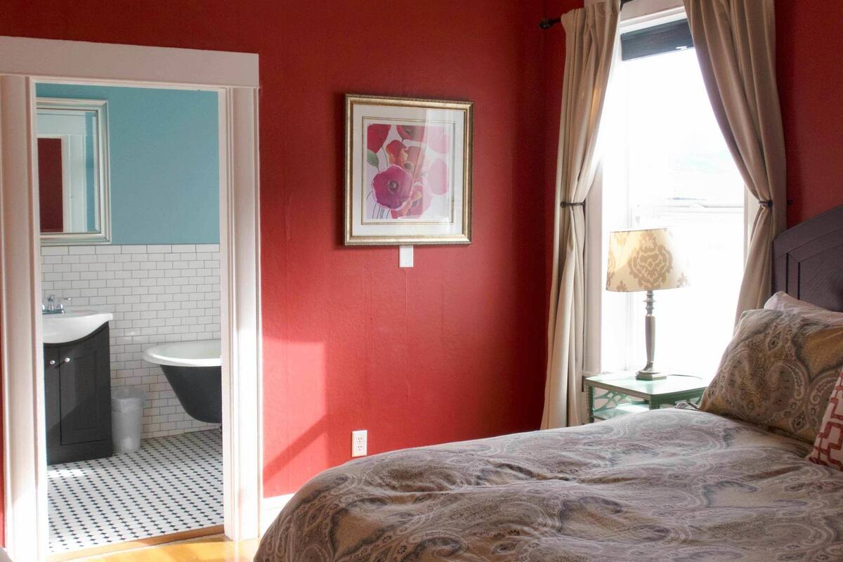 A bedroom with a bed, a ceiling fan, a window with curtains, a bedside lamp, and a framed picture on the wall.