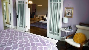 A bedroom with a bed, a chair with a yellow cushion, a small table with a lamp, and a door leading to another room. - Sauerdough Lodging (Seward)