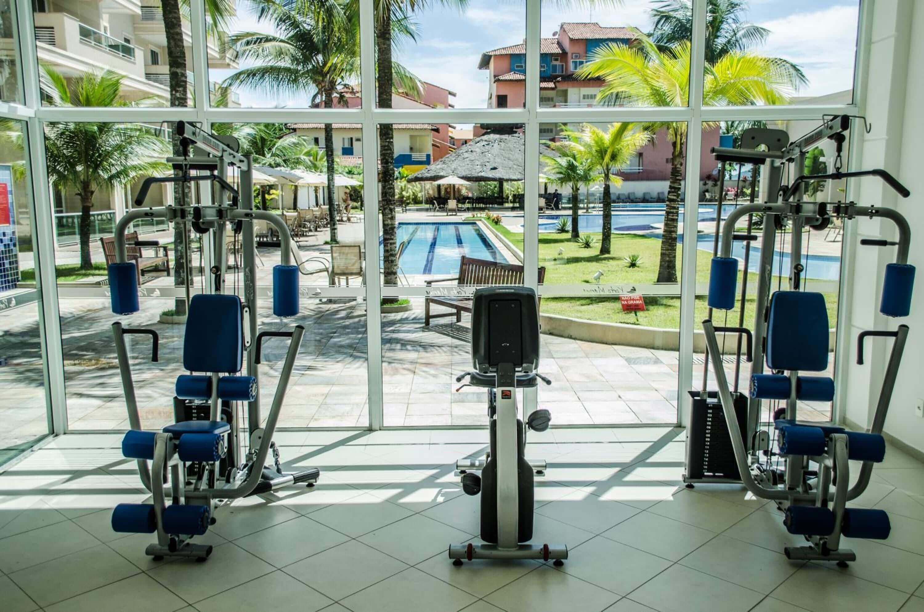 Un gimnasio con equipo de ejercicio, ventanas amplias y vistas a una piscina y palmeras.