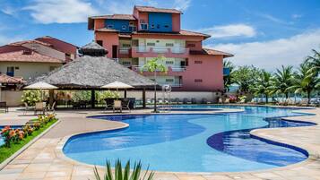Piscina com espreguiçadeiras e guarda-sóis, cercada por edifícios com varandas e um céu claro.
